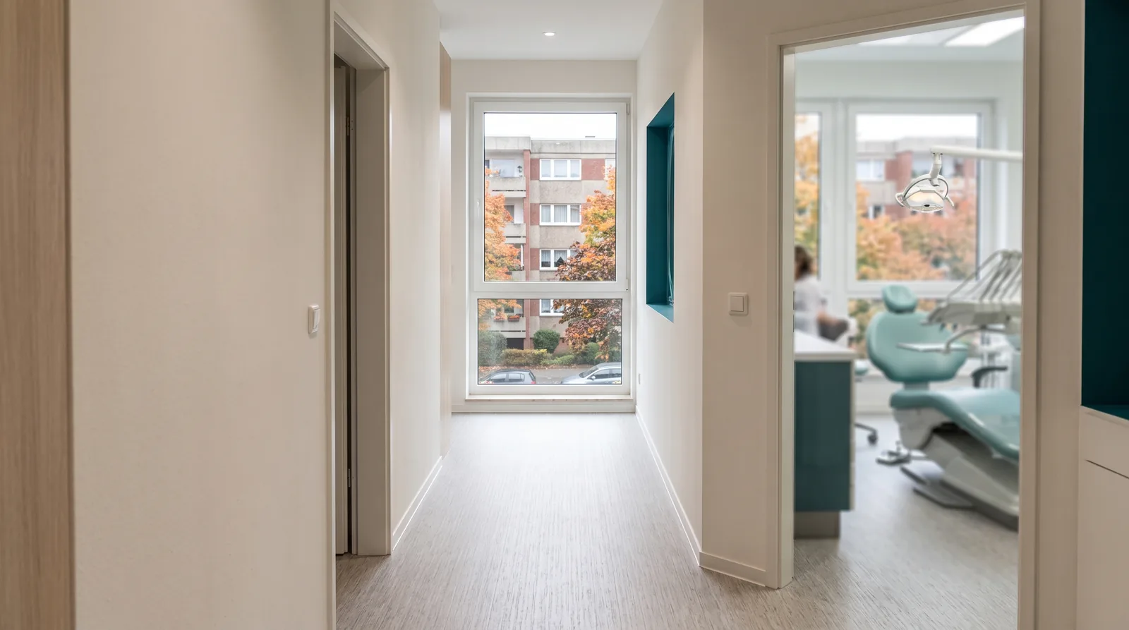Heller Praxisflur einer Zahnarztpraxis mit offener Tür zum Behandlungszimmer, durch ein Fenster Wohnblöcke aus den 1960er-Jahren und Laubbäume.